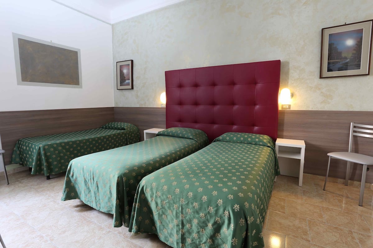 A hotel room with two single beds, a red headboard, a small table, a chair, and a wall-mounted light.