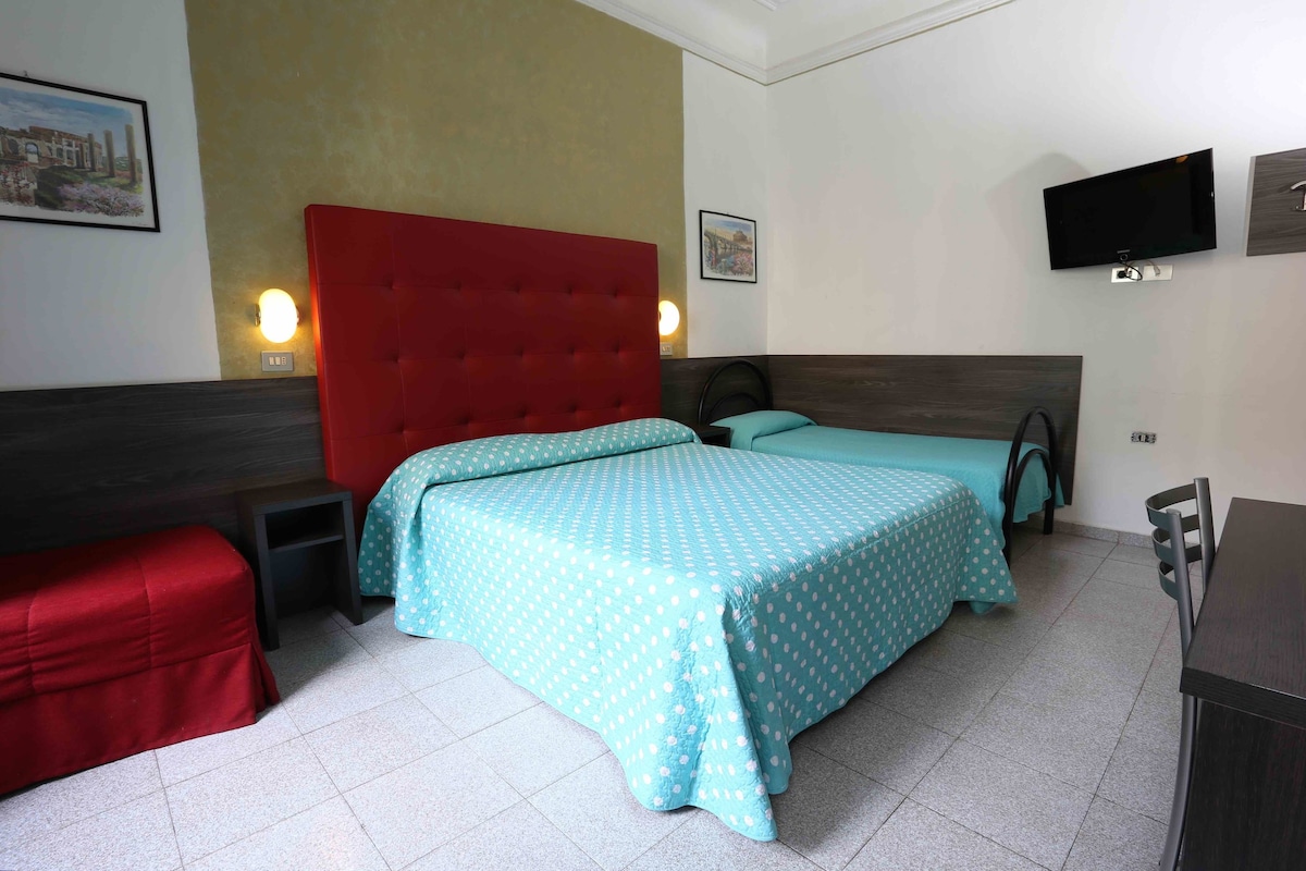 A bedroom with a red headboard, two beds with teal bedding, a TV, and a small table.