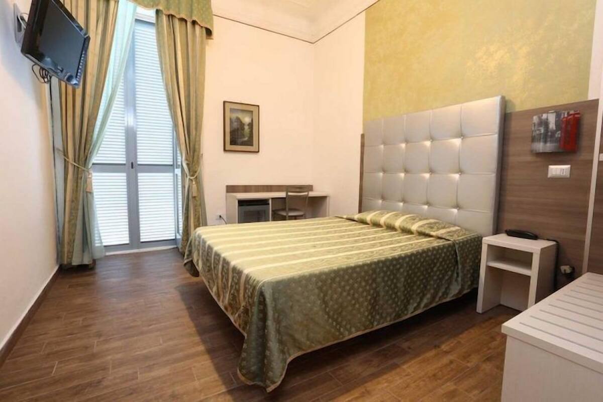 A bedroom with a bed, a desk, a chair, a television mounted on the wall, and a window with curtains.