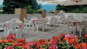 Un patio al aire libre con sillas y mesas de plástico blanco, cubierto por grandes sombrillas, rodeado de flores rojas.