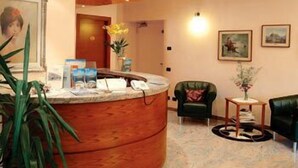 Un área de recepción con un mostrador curvo, un sillón verde, una mesita auxiliar con un florero y un cuadro en la pared.