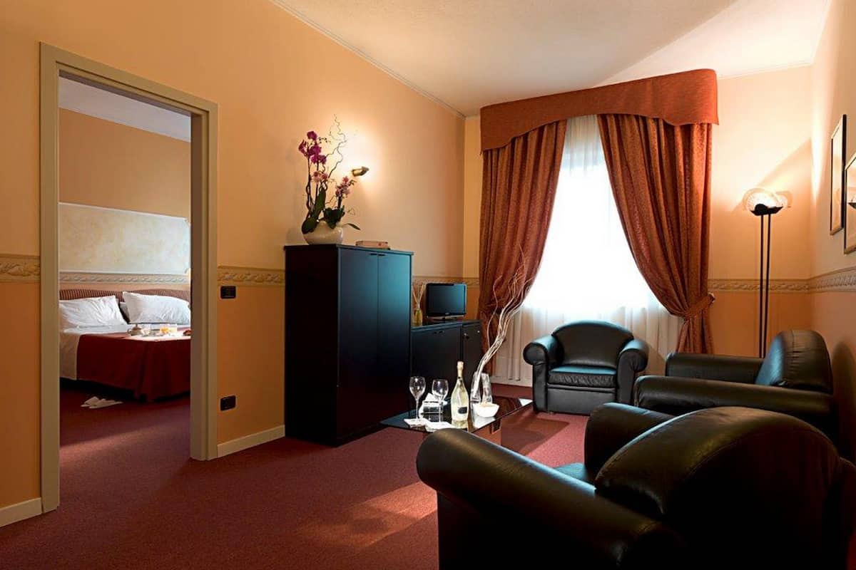 Hotelkamer met een bed, een bureau met computer, twee stoelen, een klein kastje en een raam met gordijnen.