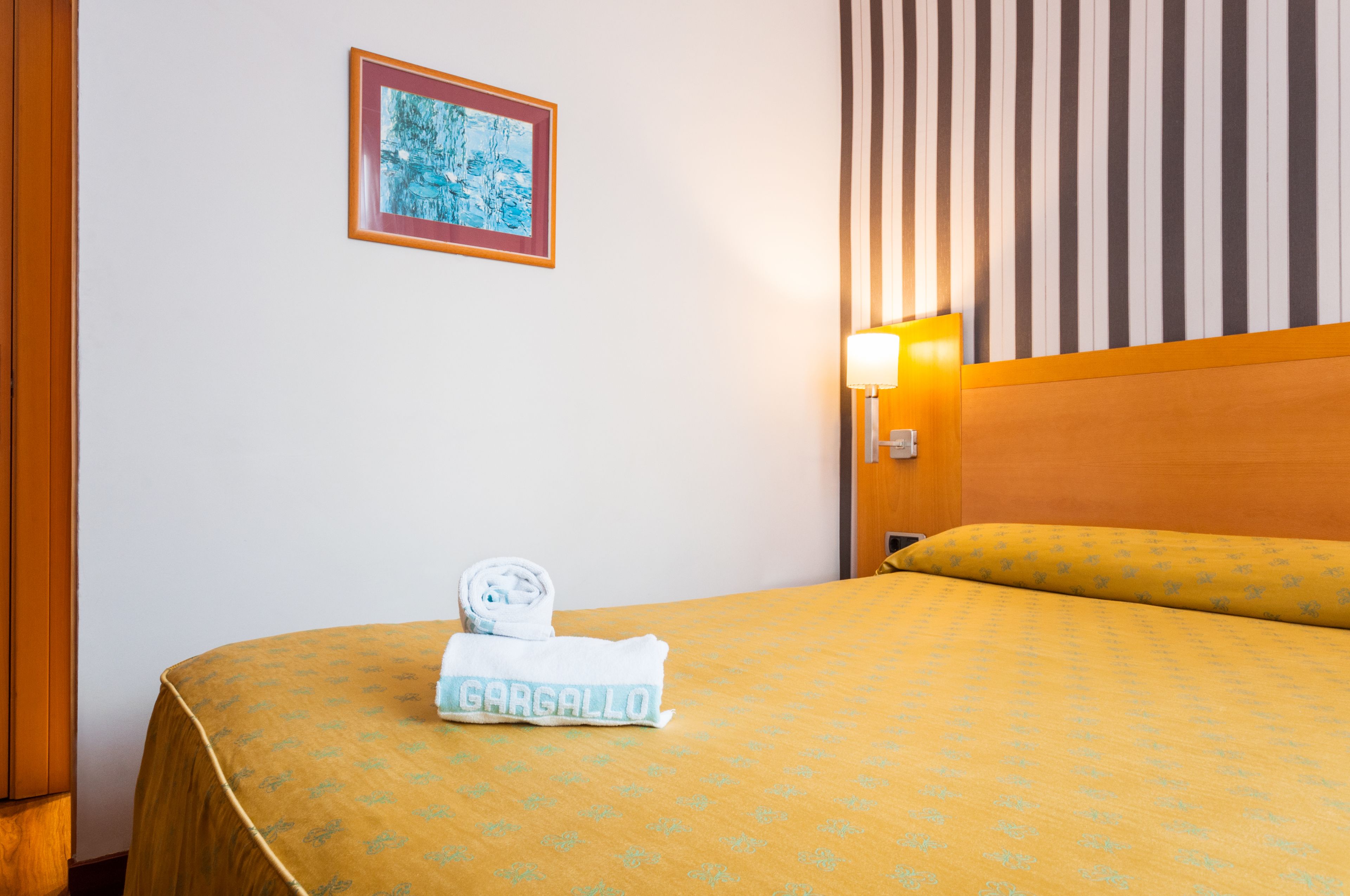 A hotel room with a bed, a framed picture on the wall, and a nightstand with a lamp.