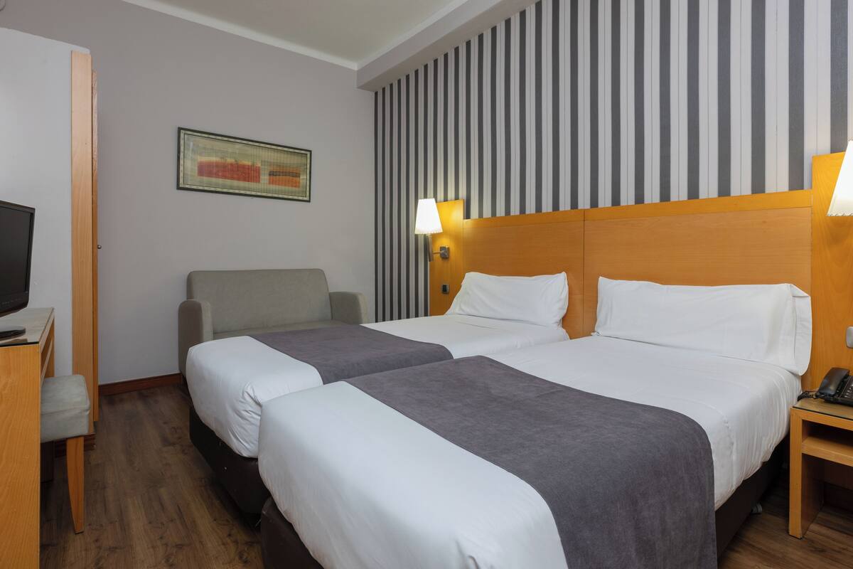 Una habitación de hotel con dos camas, un escritorio con silla, televisión y pared a rayas.