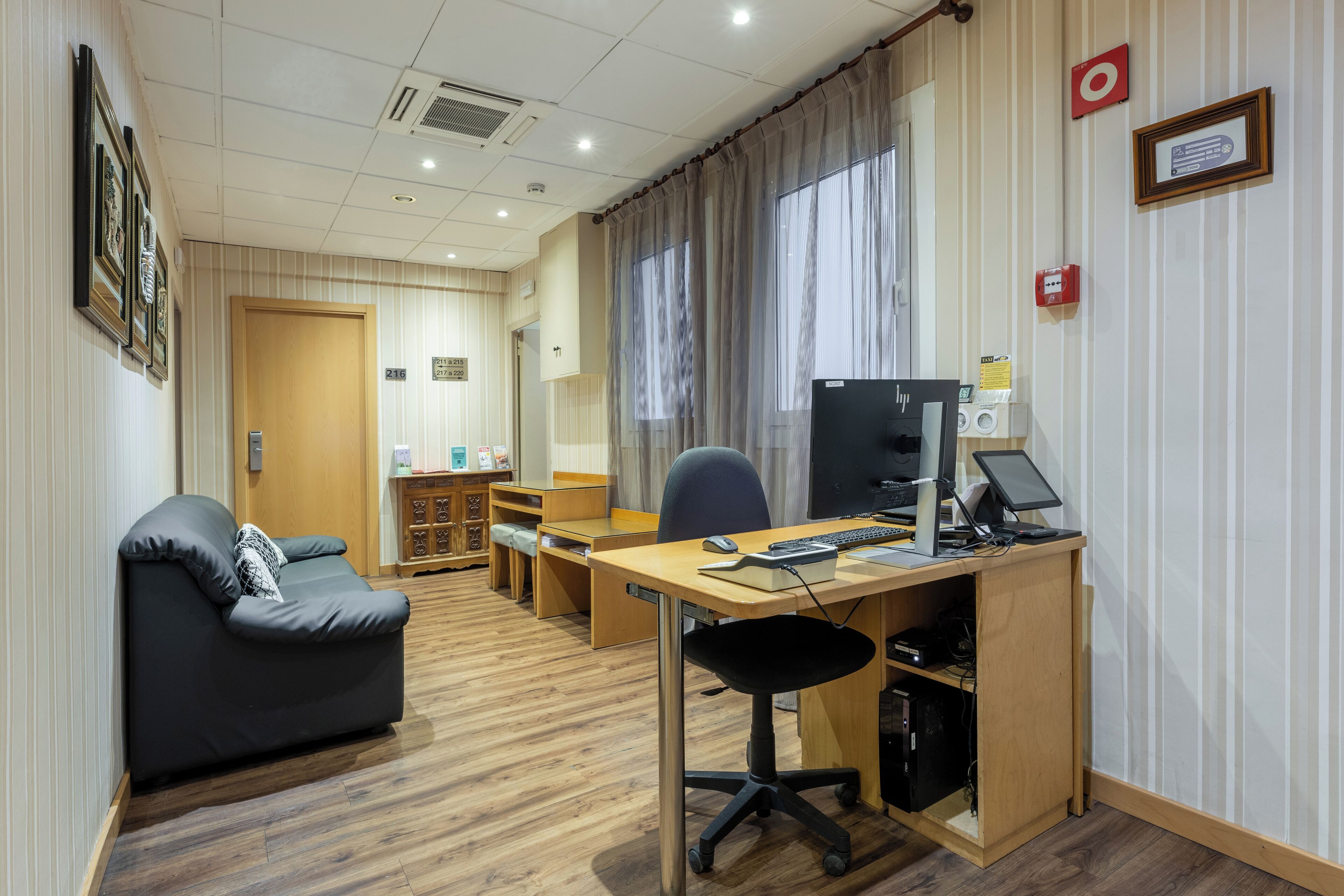 A hotel room with a wooden desk, a black office chair, a grey sofa, and a television.