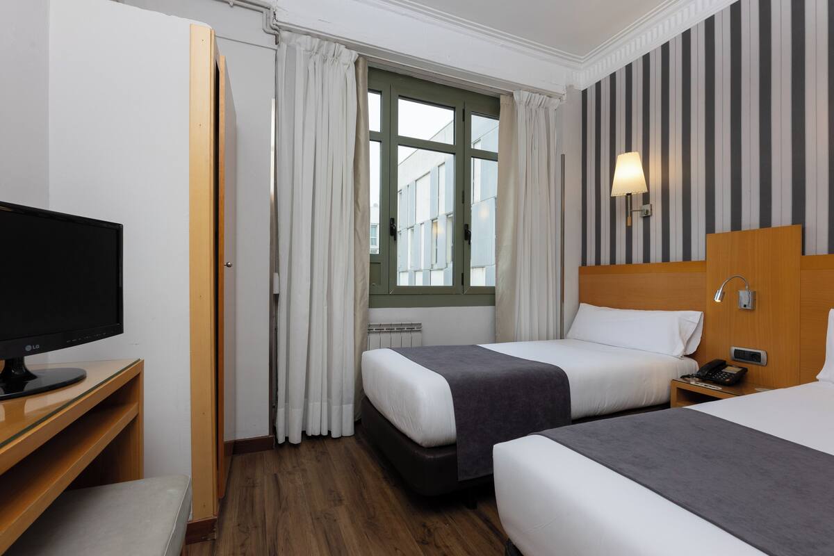 Habitación de hotel con dos camas, un escritorio con ordenador, una ventana con cortinas y una pared a rayas.