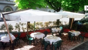 Ein Straßencafé mit Tischen und Stühlen unter weißen Sonnenschirmen, umgeben von Grünflächen und einem Backsteinpflaster.