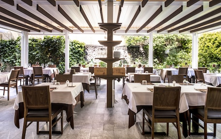 An outdoor dining area with tables and chairs, a central decorative structure, and a stone wall in the background.