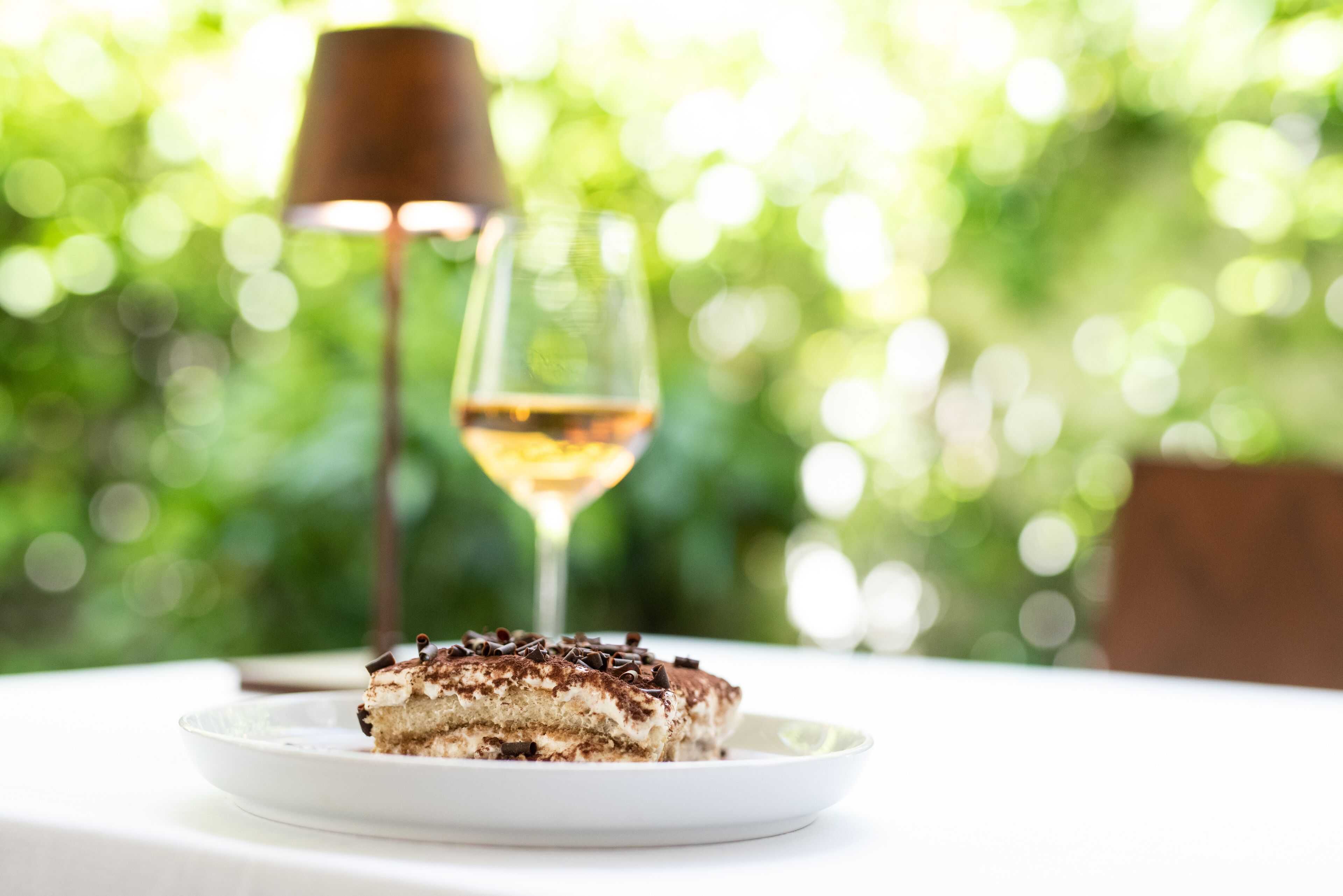 A slice of tiramisu on a white plate with a glass of white wine in the background.