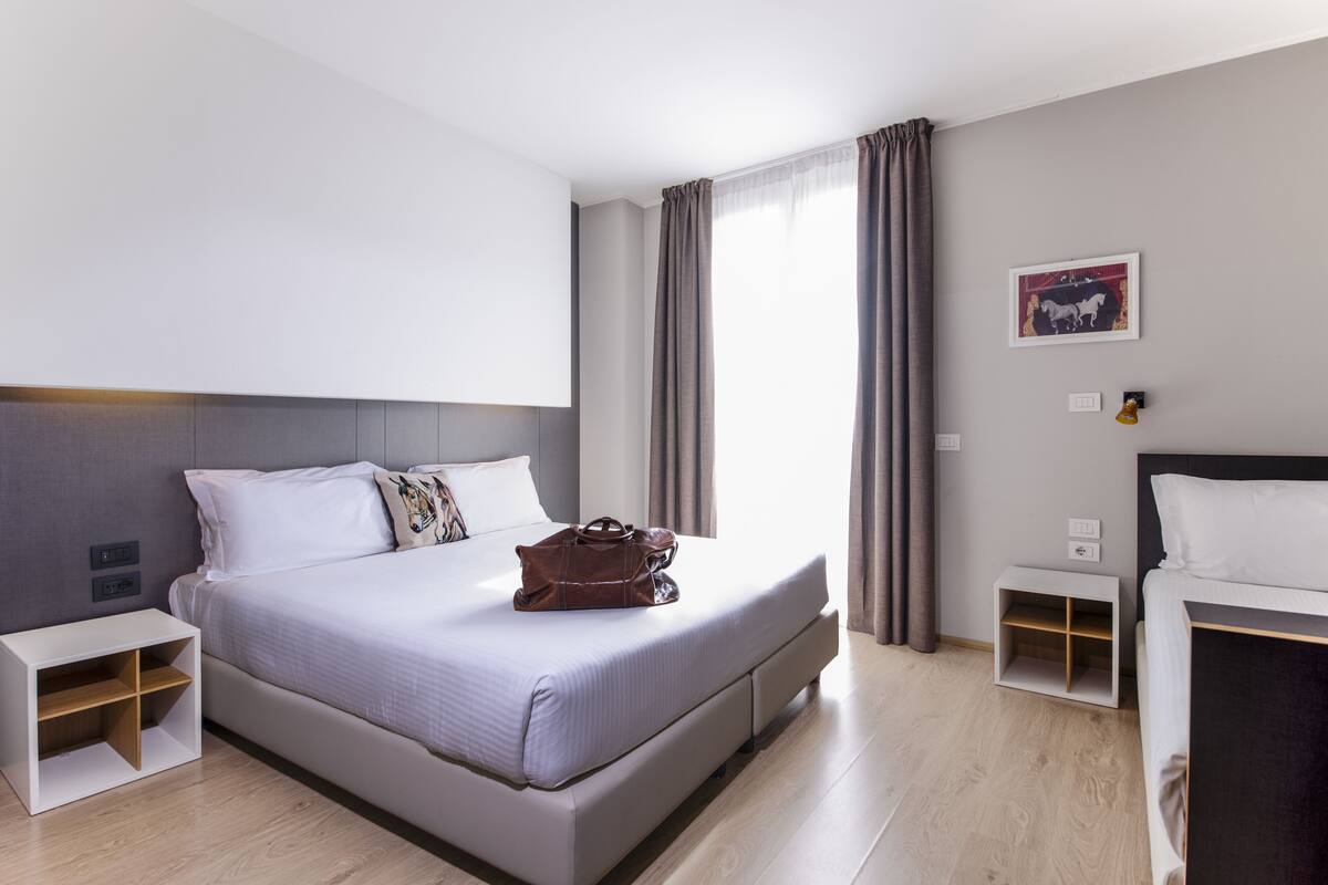 Een moderne hotelkamer met een groot bed, een nachtkastje, een klein tafeltje en een raam met gordijnen.
