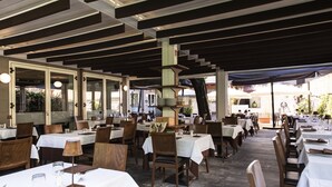 Ein Restaurant im Freien mit mehreren Tischen und Stühlen, das durch eine Dachkonstruktion überdacht ist und große Fenster, die natürliches Licht hereinlassen.