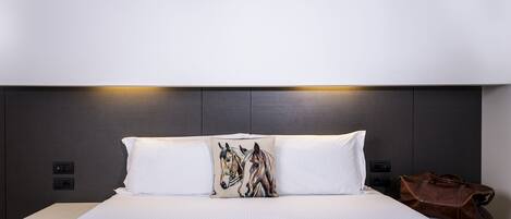 Una habitación de hotel moderna con una cama grande, ropa de cama blanca, un cabecero oscuro y un bolso de cuero marrón sobre la mesita de noche.