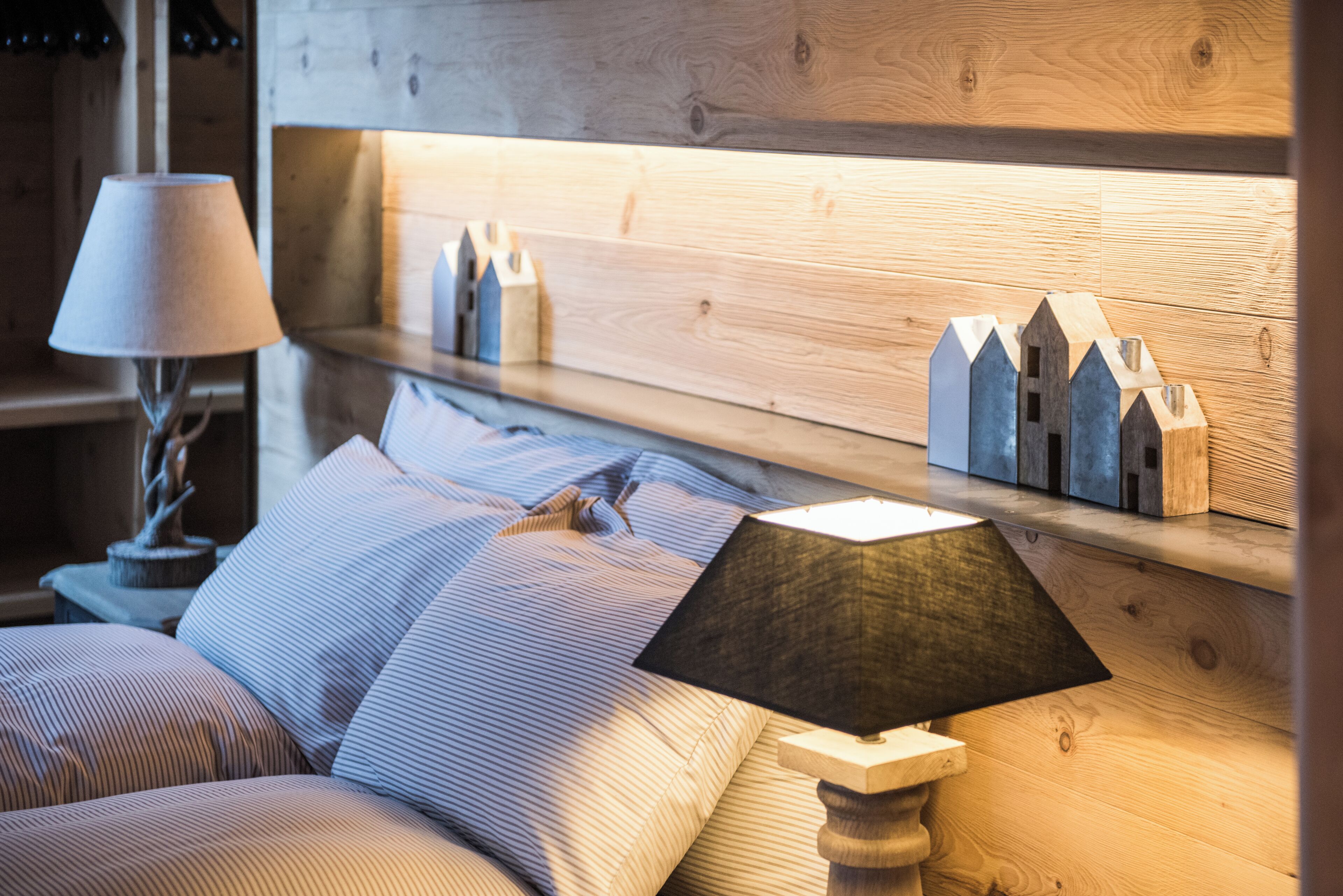Un dormitorio con cabecera de madera, lámpara de noche, cama con ropa de cama a rayas y estante con adornos.