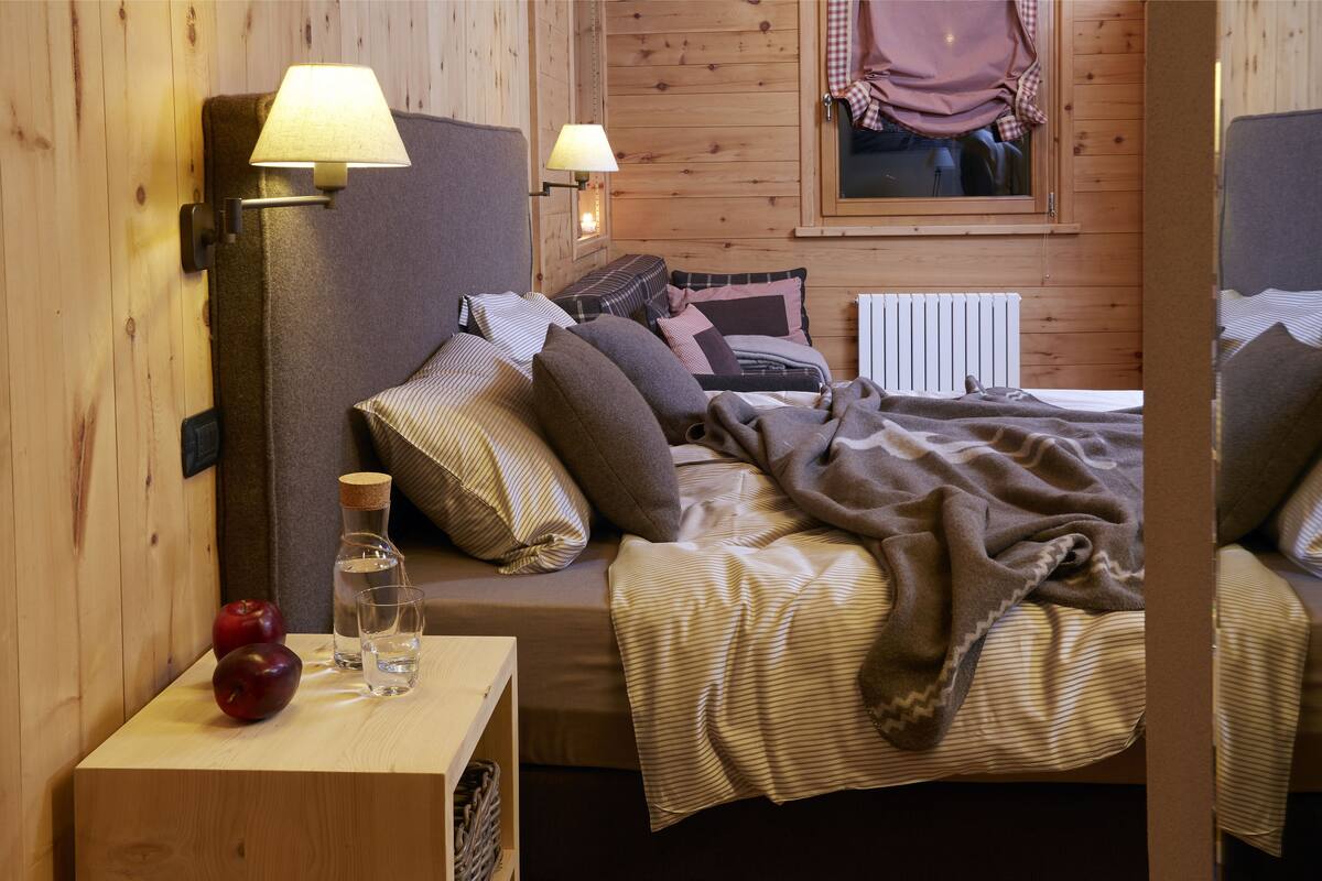 A cozy wooden room with a bed, pillows, a blanket, a nightstand with apples and a glass of water, and two wall-mounted lamps.