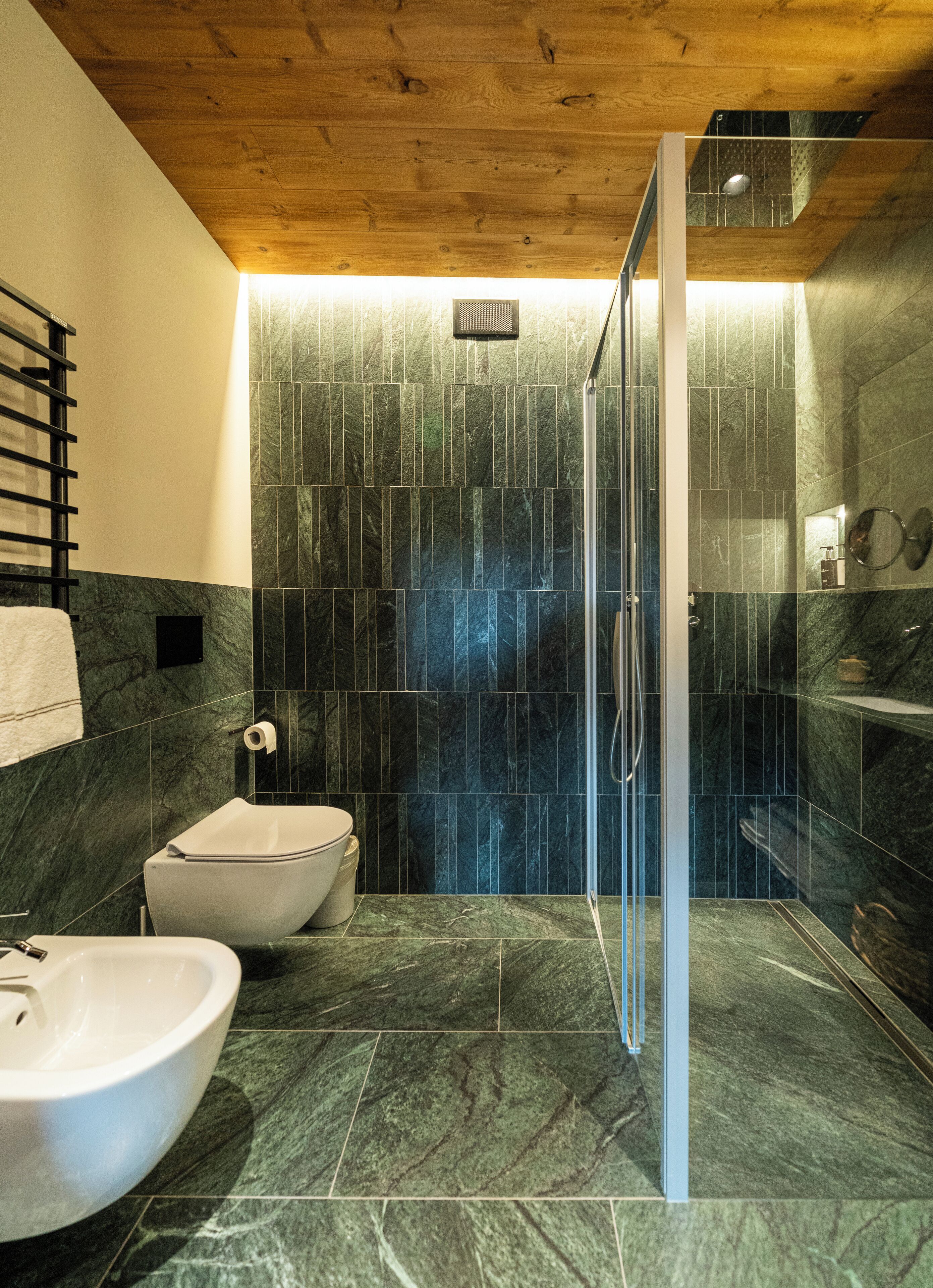 A modern bathroom with a wooden ceiling, a glass shower enclosure, a white sink, and a bidet.