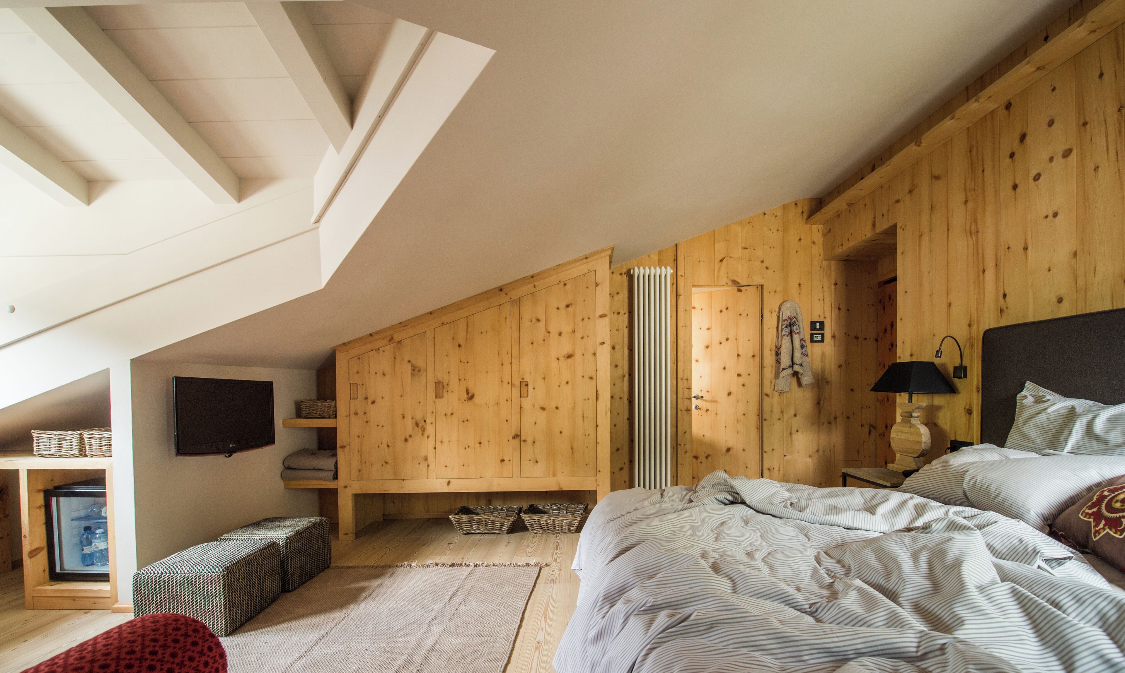 A bedroom with a sloped ceiling, wooden walls, a bed with striped bedding, a bedside table with a lamp, and a television on the wall.