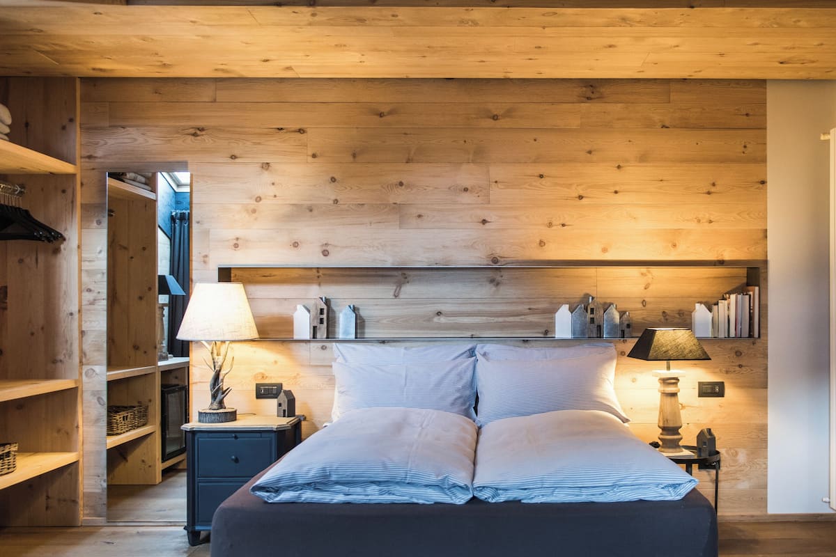 A bedroom with a wooden ceiling, a bed with striped bedding, two bedside tables with lamps, and a shelf with books and decorative items.