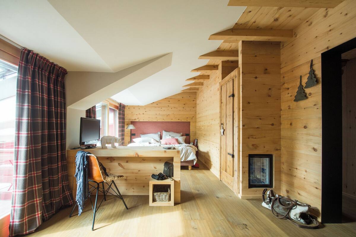A cozy cabin bedroom with a wooden ceiling, a bed with pillows, a desk with a chair, and a window with curtains.