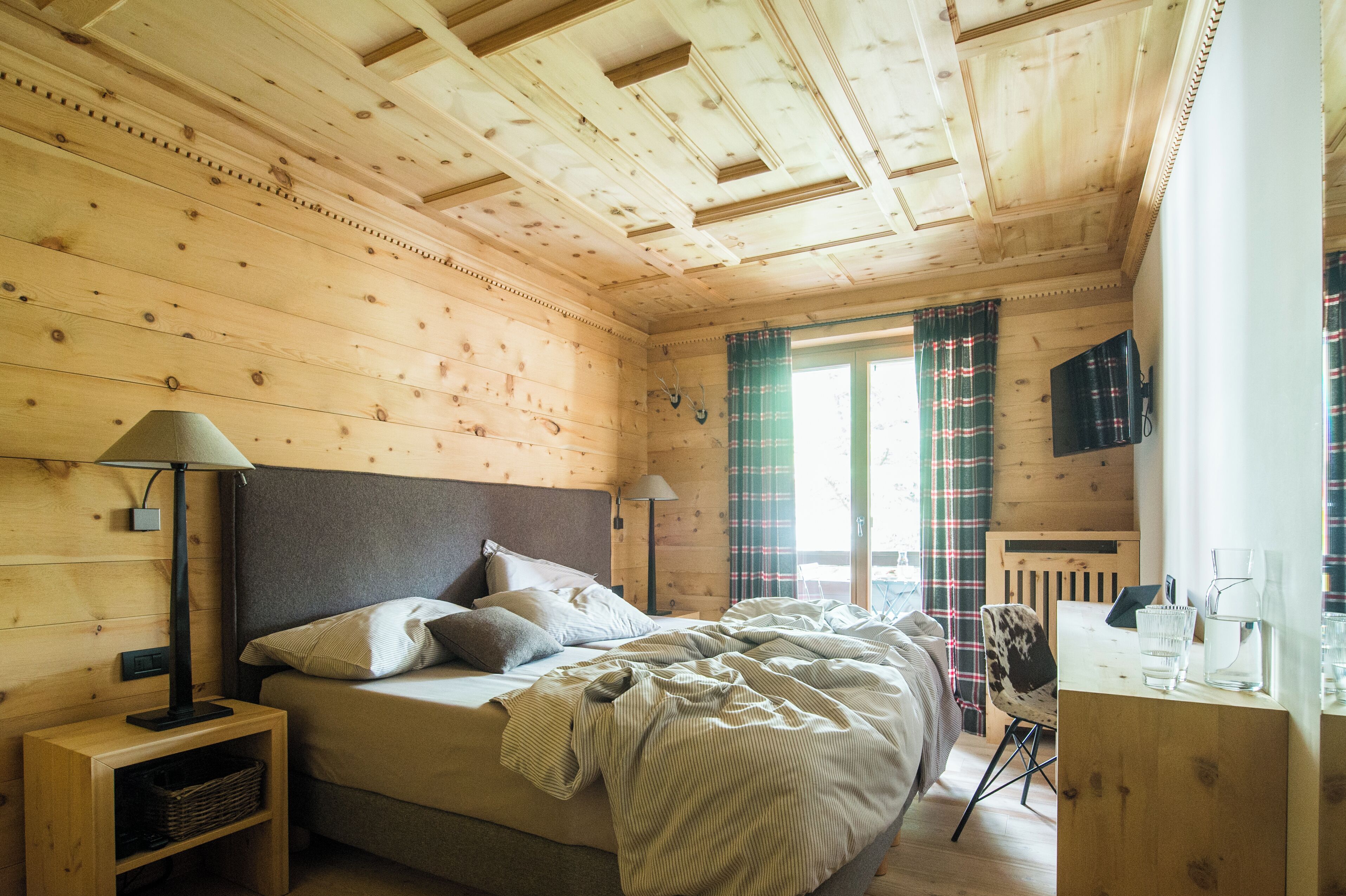 A wooden cabin bedroom with a bed, nightstands, a desk, and a window with curtains.
