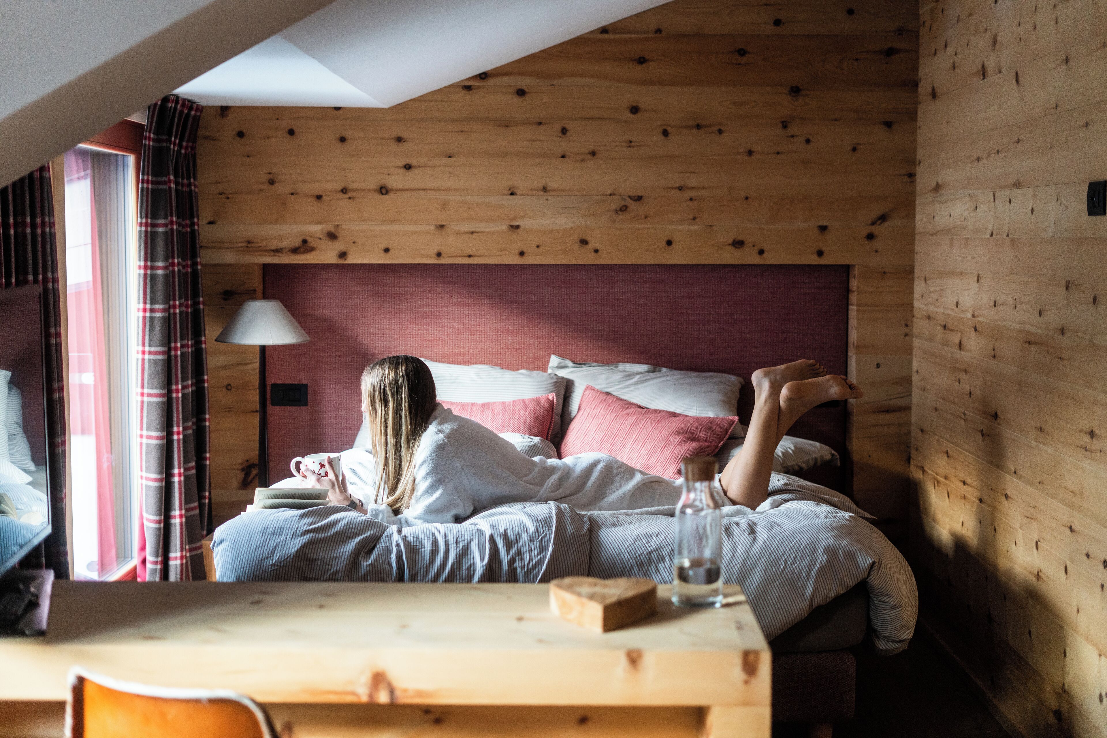 A cozy room with a wooden ceiling, a bed with red and white bedding, a bedside table with a lamp, and a window with curtains.