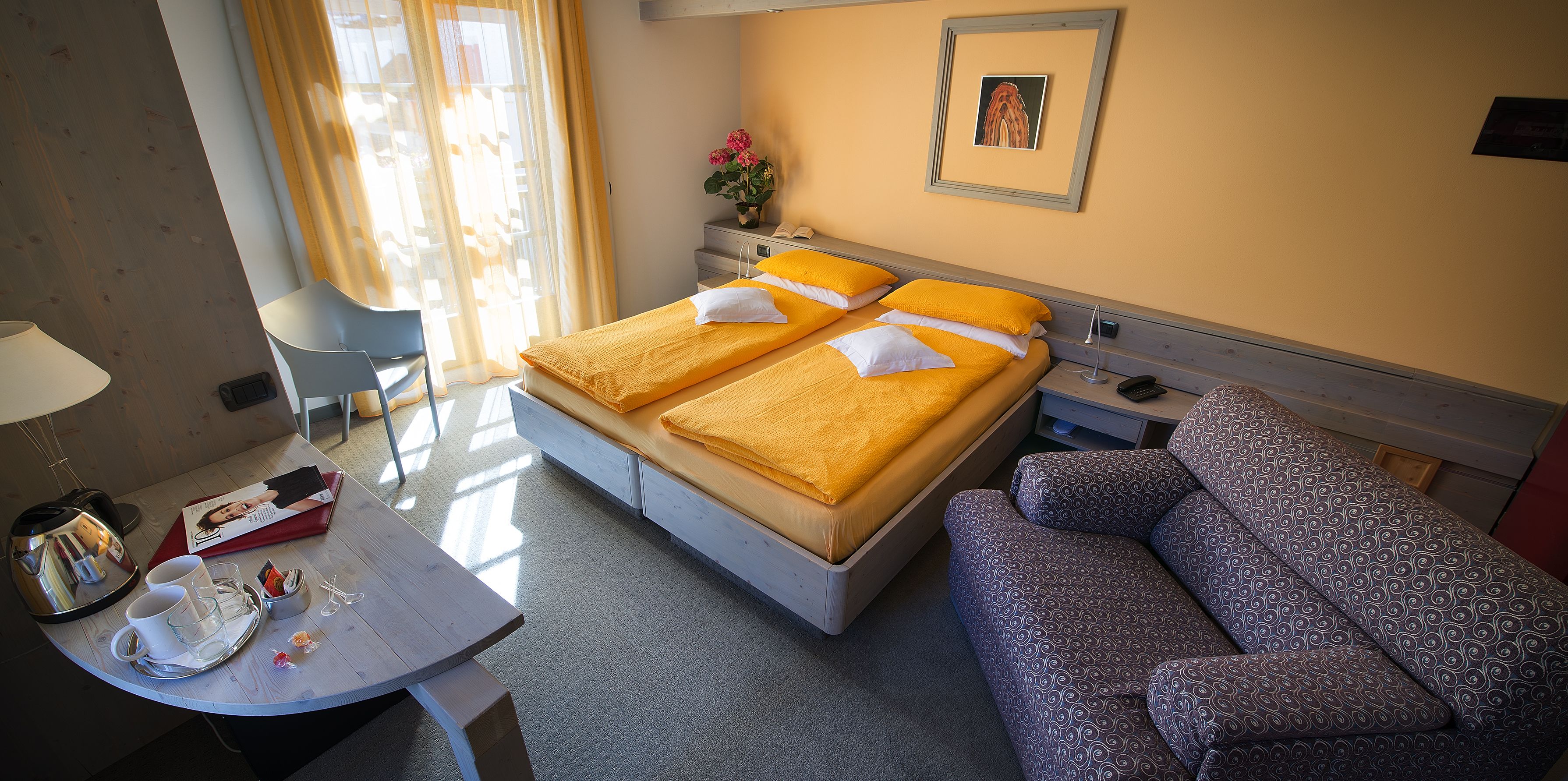 A hotel room with a bed, a sofa, a desk with a kettle, cups, and a magazine.