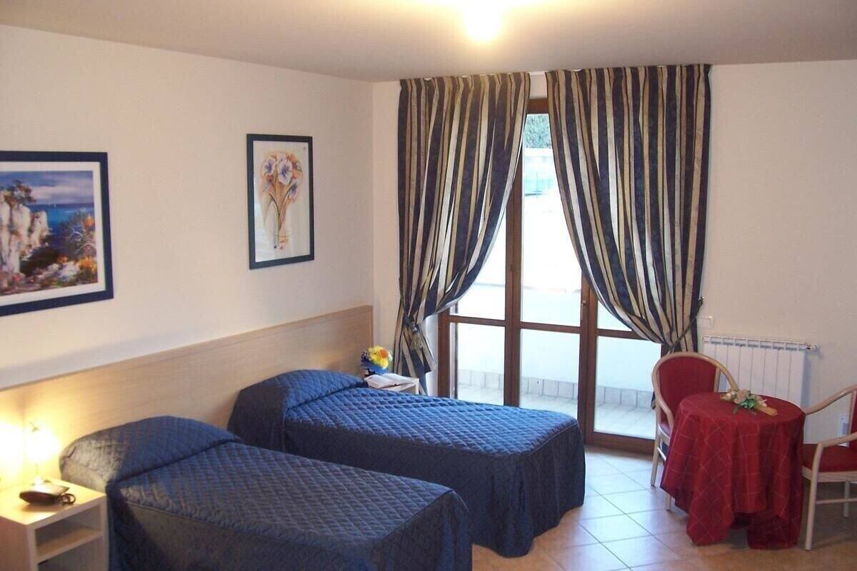 A room with two beds, a small table with a red tablecloth, and a chair. There is a window with curtains, a radiator, and a picture on the wall.