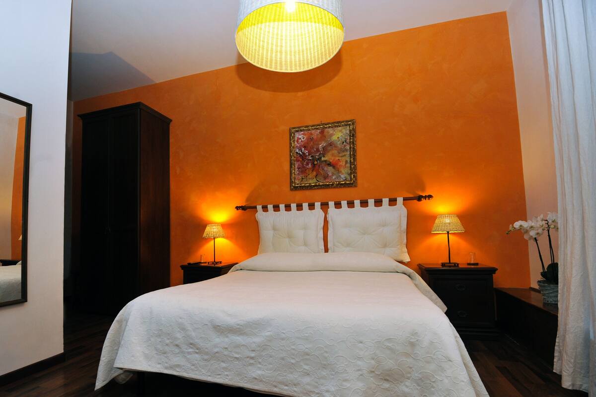 A hotel room with a large bed, two bedside tables with lamps, a painting on the wall, and a mirror on the left.
