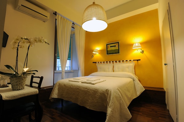 Una cama bien hecha con una colcha blanca, una lámpara de noche y un cuadro en la pared. Hay una ventana con cortinas blancas, una planta en maceta y una silla con una mesita de noche.