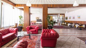 A spacious lobby with red leather sofas, a glass coffee table, and a patterned rug. There are potted plants and a wooden ceiling with a geometric design.