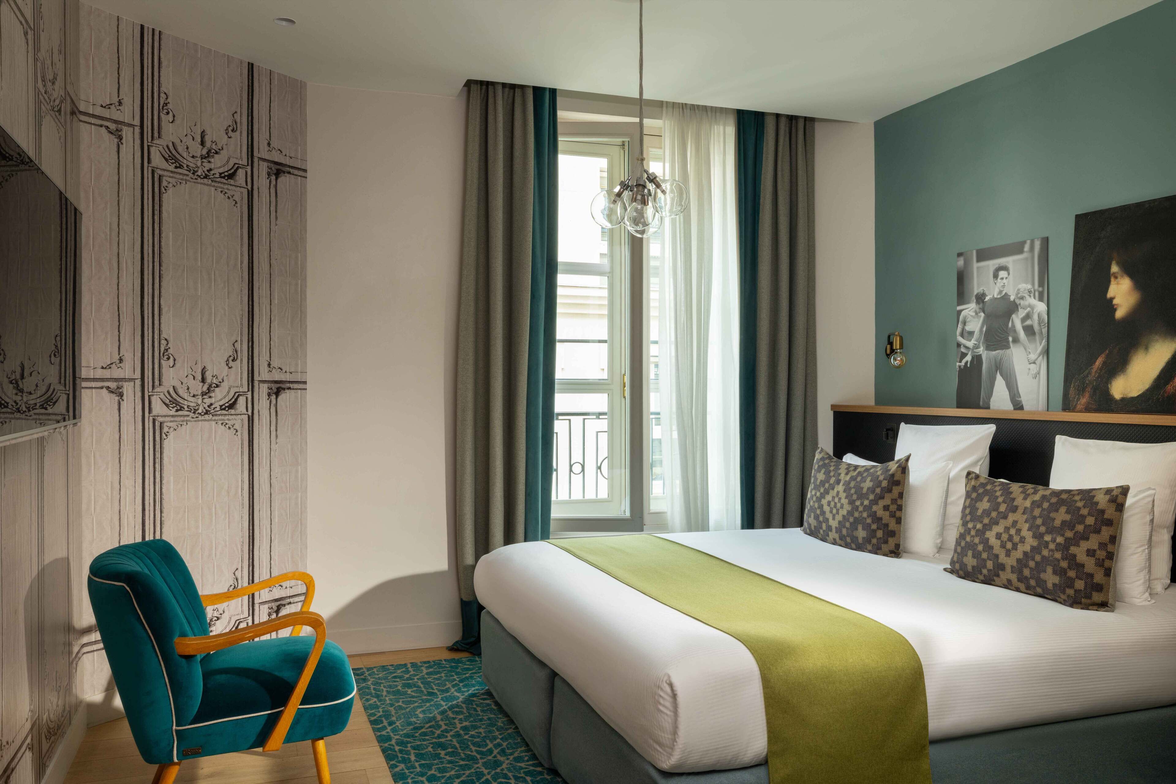 A hotel room with a bed, a chair, a window with curtains, and a painting on the wall.