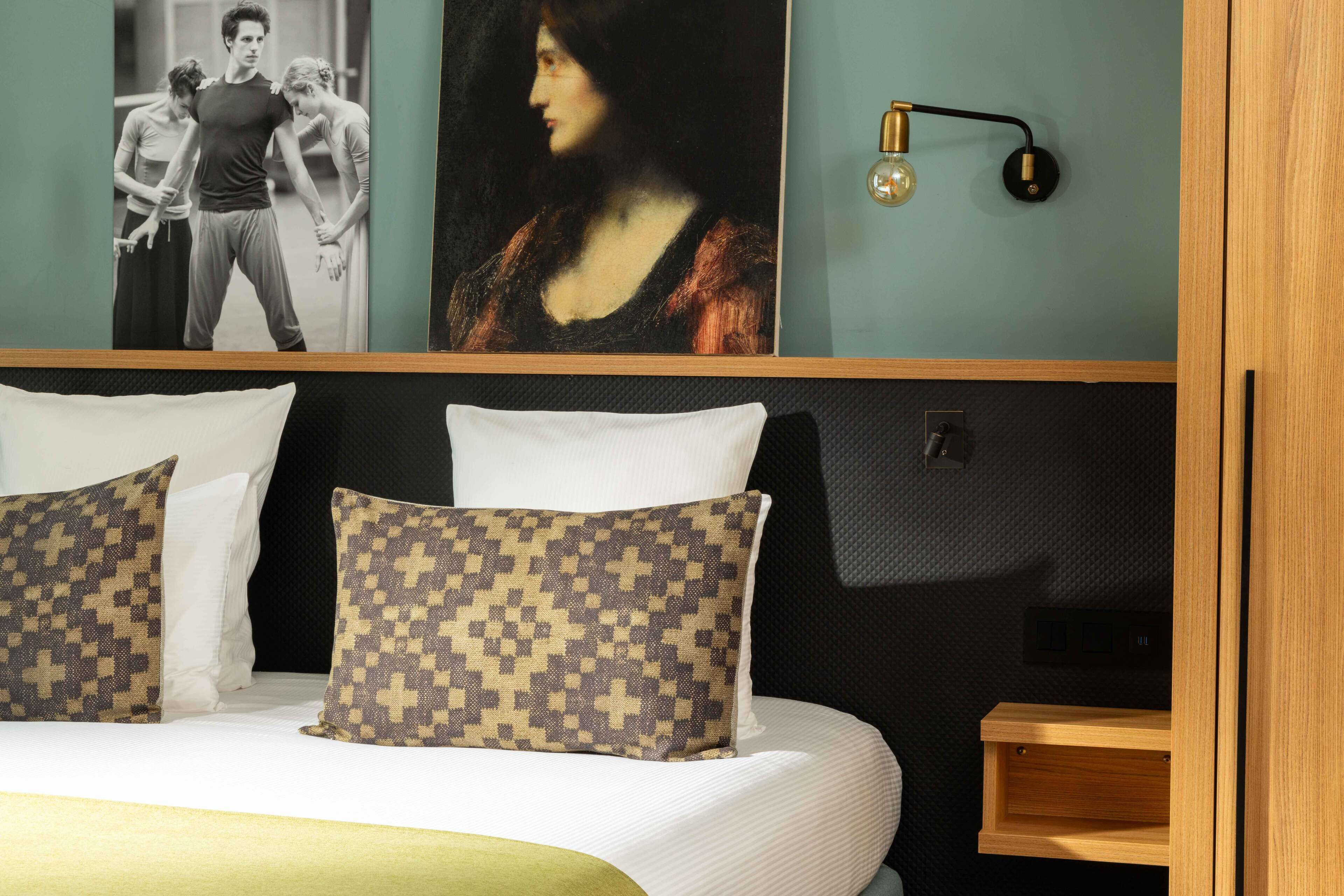 Una cama bien hecha con almohadas estampadas, una lámpara de pared y una obra de arte enmarcada sobre la cama.