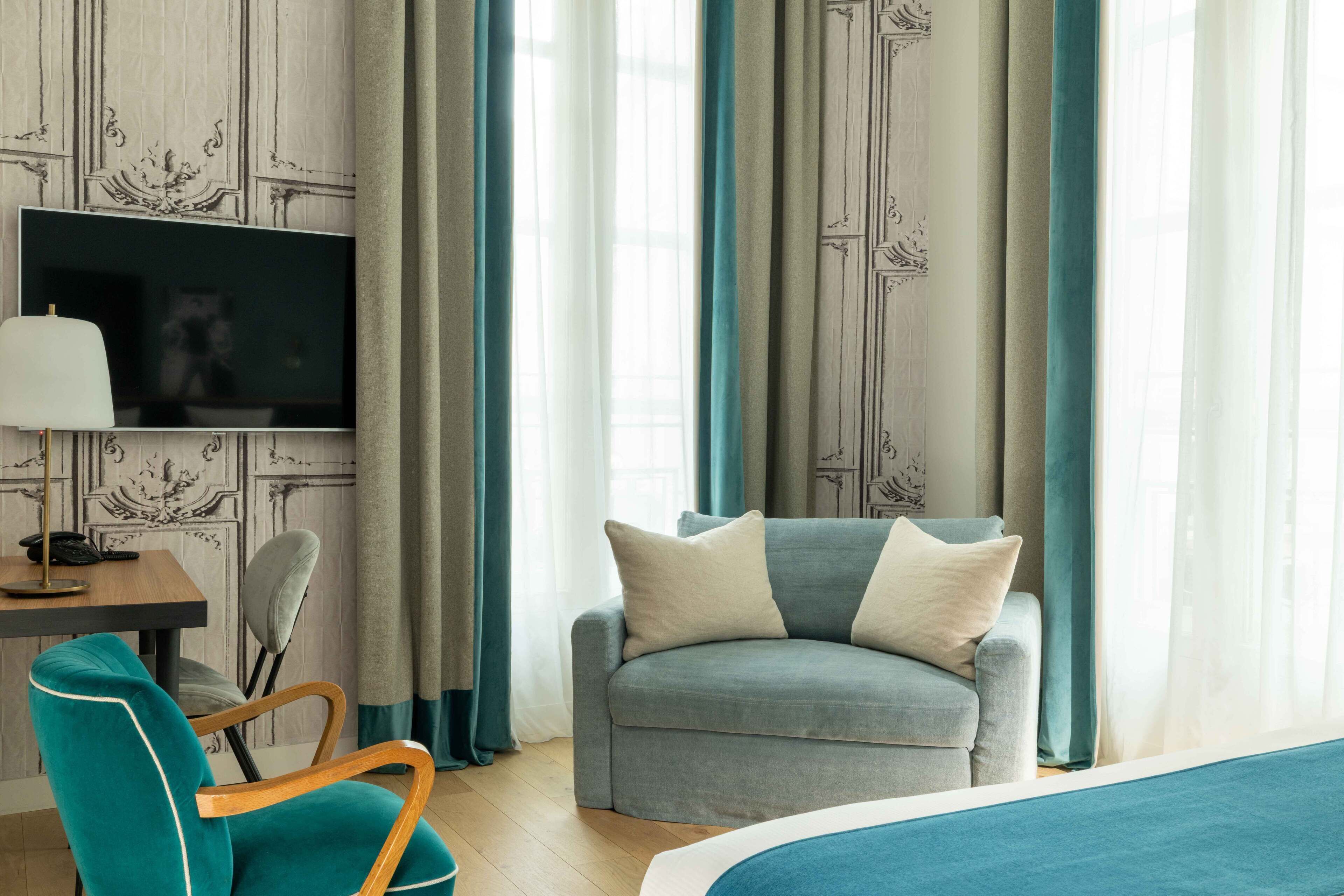 A hotel room with a sofa, a TV, a desk with a lamp, and curtains.