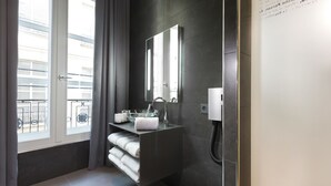 Et moderne badeværelse med væg af mørke fliser, vask af glas, et spejl og et vindue med gardiner.