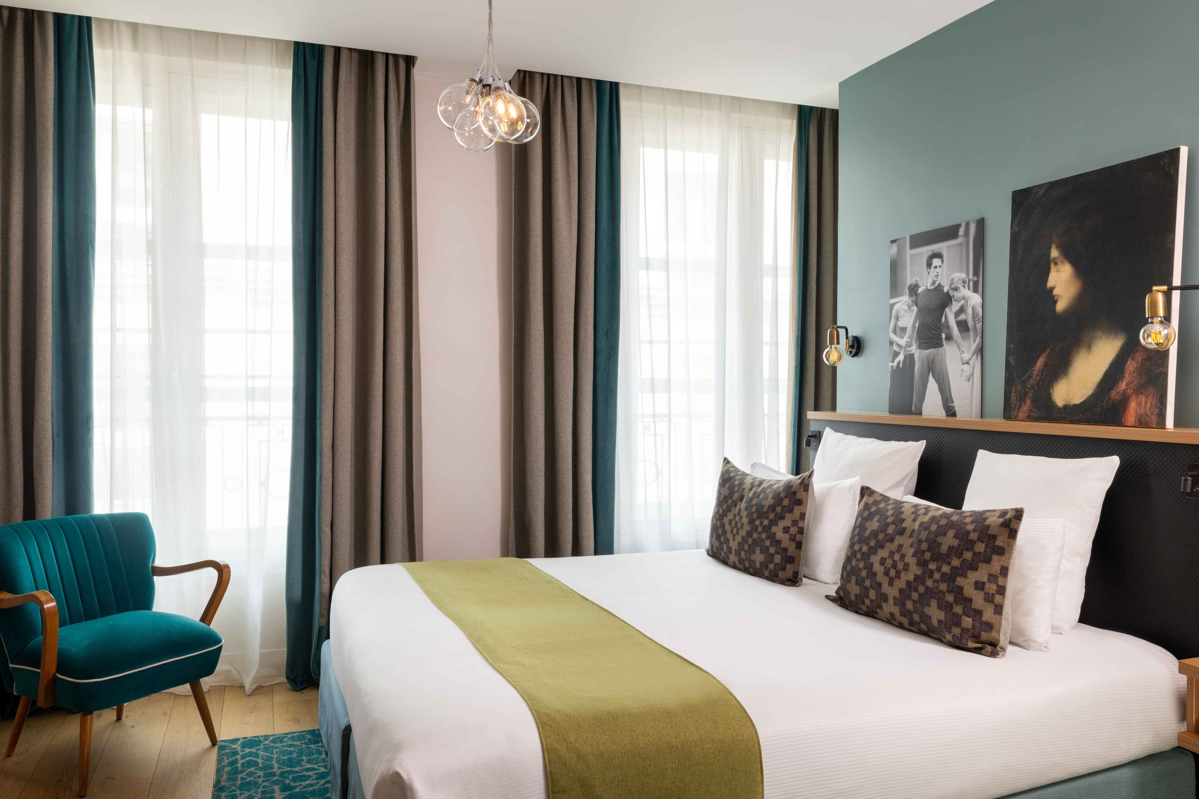 A hotel room with a large bed, a teal armchair, and a view of the city through sheer curtains.