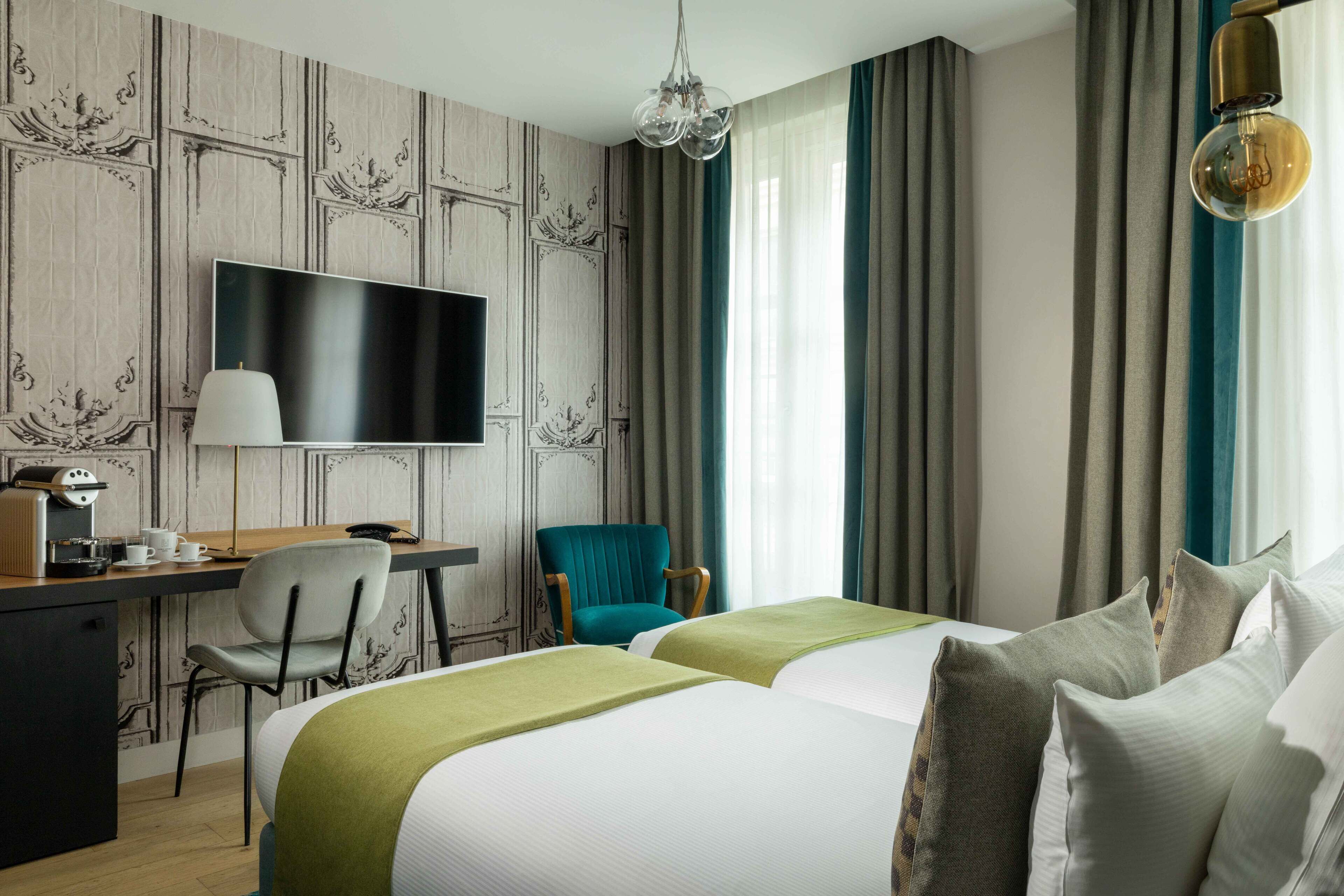 Une chambre d’hôtel équipée d’une télévision à écran plat, d’un bureau avec une machine à café, d’un fauteuil turquoise, de deux lits avec une literie verte et blanche, et d’un lustre.