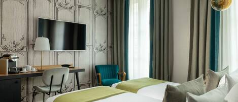 Une chambre d’hôtel équipée d’une télévision à écran plat, d’un bureau avec une machine à café, d’un fauteuil turquoise, de deux lits avec une literie verte et blanche, et d’un lustre.