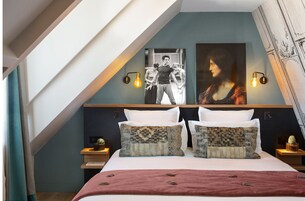 A bedroom with a sloped ceiling, a bed with patterned pillows, two wall-mounted light fixtures, and two paintings.
