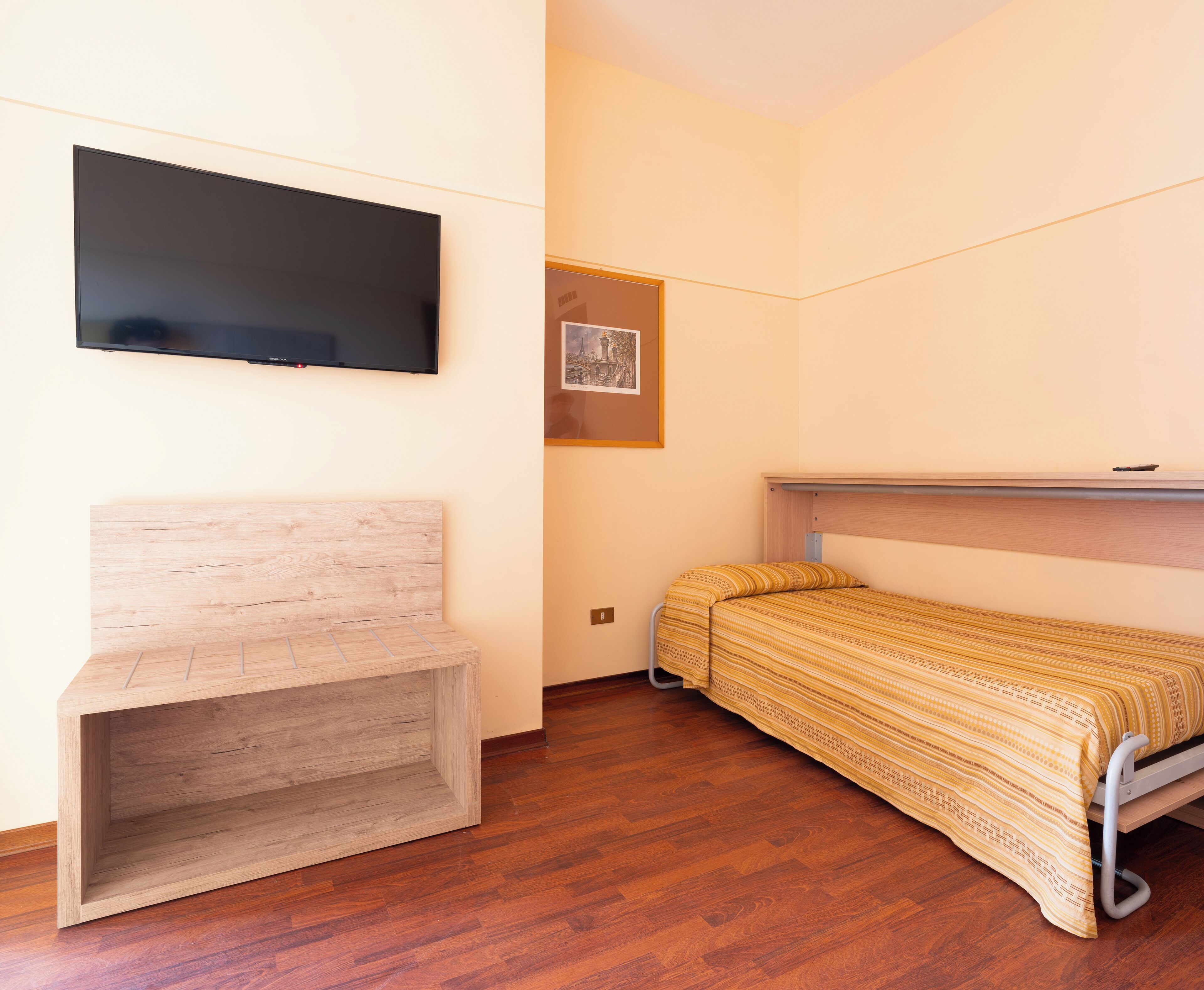 A small, well-lit room with a wooden bench, a bed with a patterned bedspread, a mounted flat-screen TV, and a framed picture on the wall.