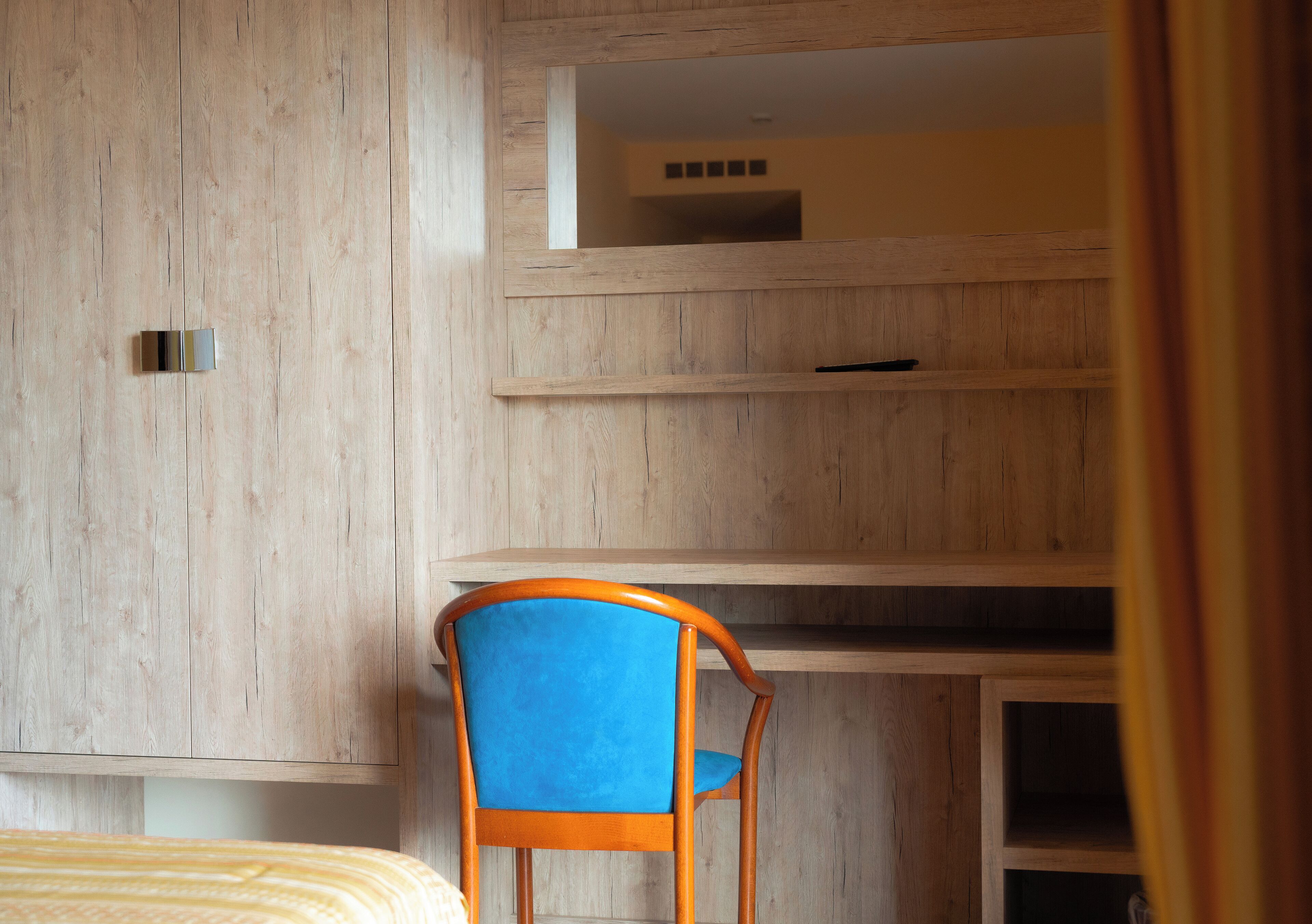 A room with a bed, a wooden wardrobe, a blue chair, and a desk with shelves.