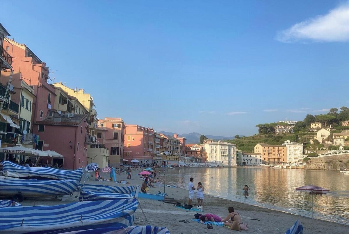 A beach with striped umbrellas and boats, people relaxing, and buildings along the water.