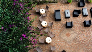 Un patio al aire libre con mesas y sillas redondas, rodeado de piso de ladrillo y abundante vegetación.