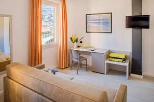 A modern hotel room with a sofa, a desk, a chair, a TV, and a window with curtains.