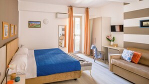 Habitación de hotel con cama, sofá, escritorio y silla. Cuenta con una ventana con cortinas, un televisor de pantalla plana y cuadros en la pared.