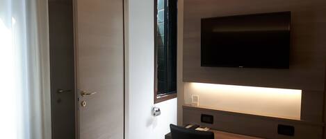 A hotel room with a closed door, a flat-screen TV mounted on the wall, a bedside table, and a chair.