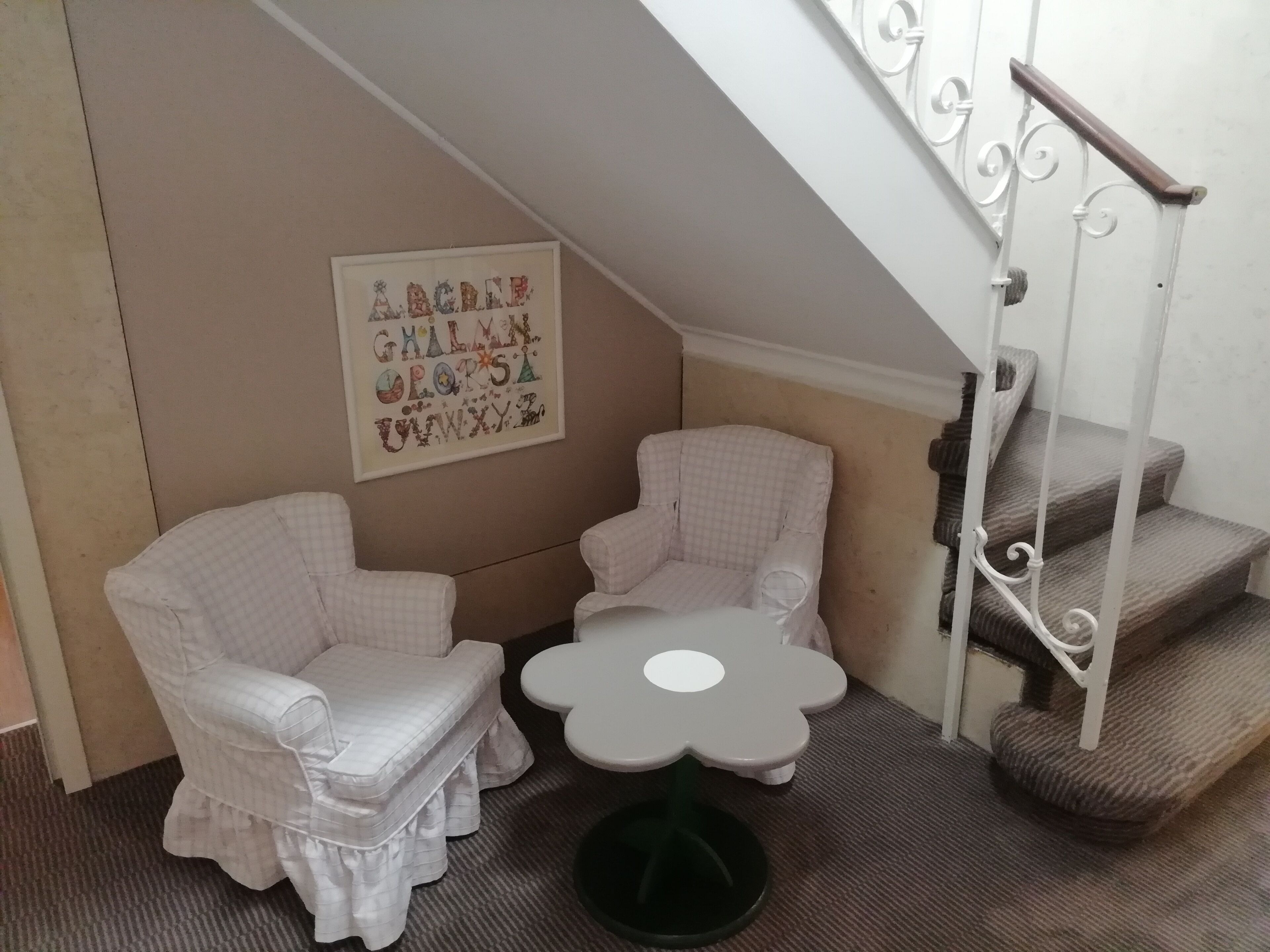A corner room with two white armchairs, a small table, a staircase, and a wall with alphabet letters.