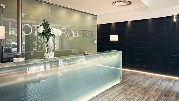 A reception area with a glass counter, a flower vase, and a dark tiled wall. - Hotel Roberta (Mestre)