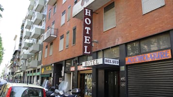 Un hôtel avec une enseigne indiquant qu’il s’agit bien d’un hôtel, un commerce dont la devanture est fermée et voletée, et un scooter garé.
