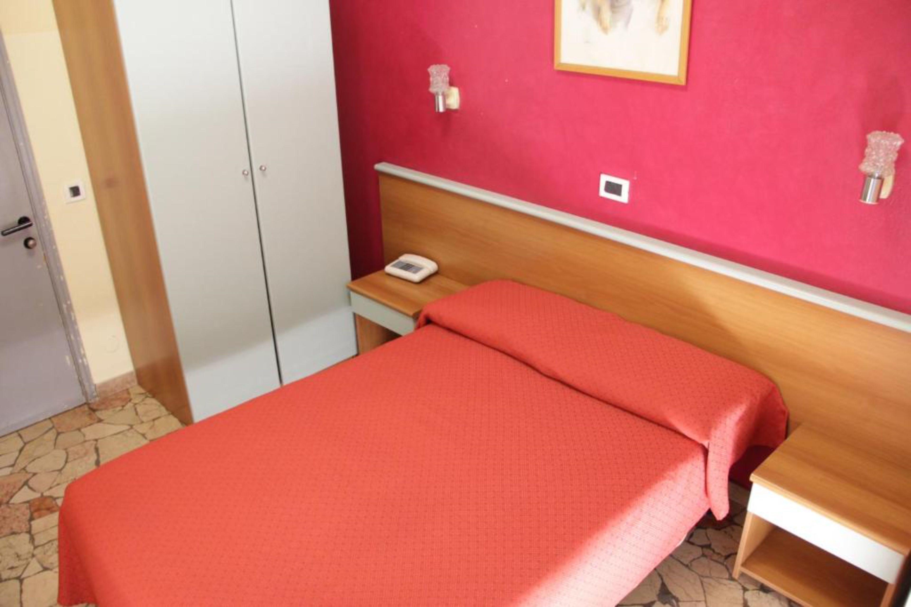 A single bed with a red bedspread in a room with a red wall, a wooden headboard, a bedside table with a lamp, and a white wardrobe.