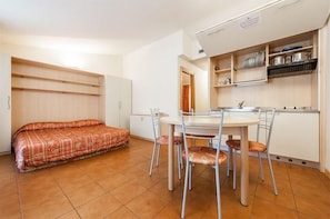 A compact room with a bed, a dining table with chairs, a kitchen area with shelves, and a white cabinet. - Club Hotel la Vela (Nago-Torbole)