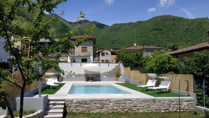 Un espace piscine agrémenté d’un mur en pierre, de chaises longues blanches et offrant une vue sur des collines verdoyantes et des bâtiments en arrière-plan.